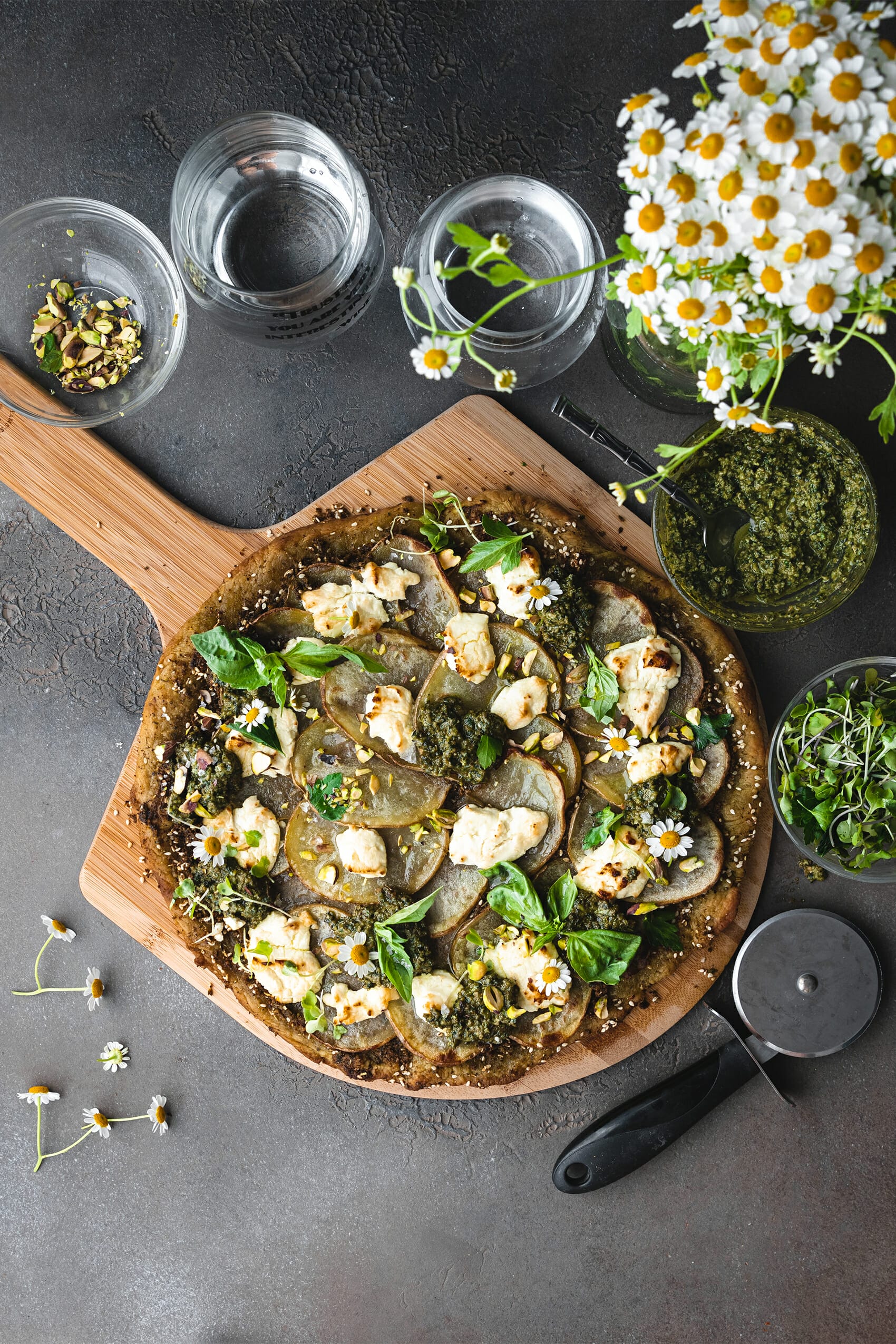 overhead shot of vegan pesto potato pizza on a pizza peel next to a bowl of pesto and some fresh herbs