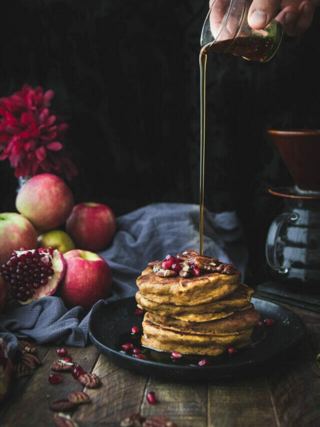 hand pouring maple syrup onto a stack of vegan pumpkin pancakes