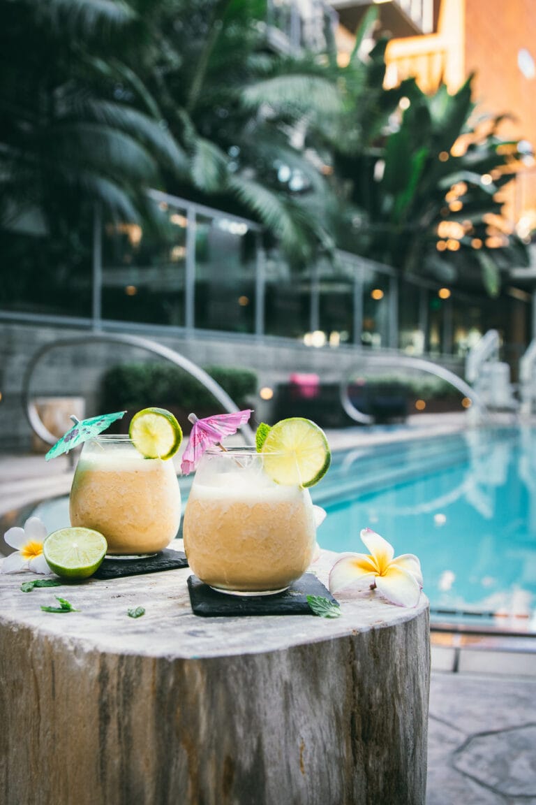 two 4 ingredient pina coladas next to a pool on a wooden stand