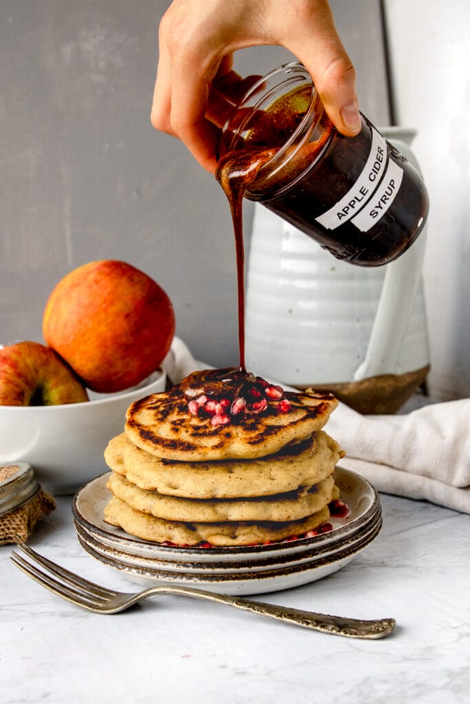 hand holding jar of boiled apple cider pouring it onto a stack of pancakes