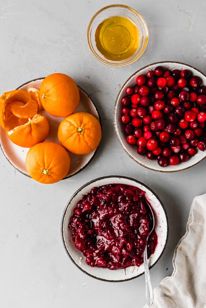 orange cranberry sauce in a white bowl next to bowls of fresh cranberries and maple syrup