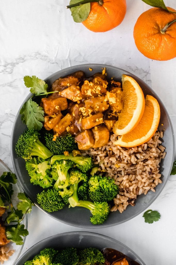 overhead view of vegan orange tofu with brown rice and broccoli on a round black plate