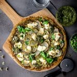 overhead shot of vegan pesto potato pizza on a pizza peel next to a bowl of pesto and some fresh herbs