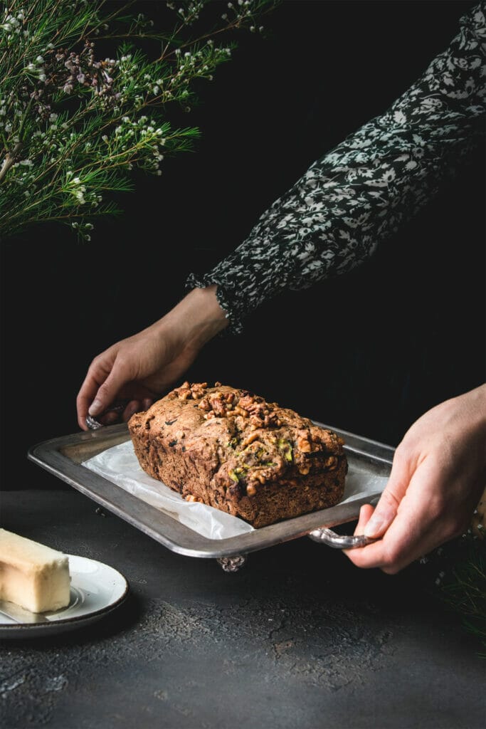 Two hands holding a silver tray with a loaf of plant-based zucchini bread on it