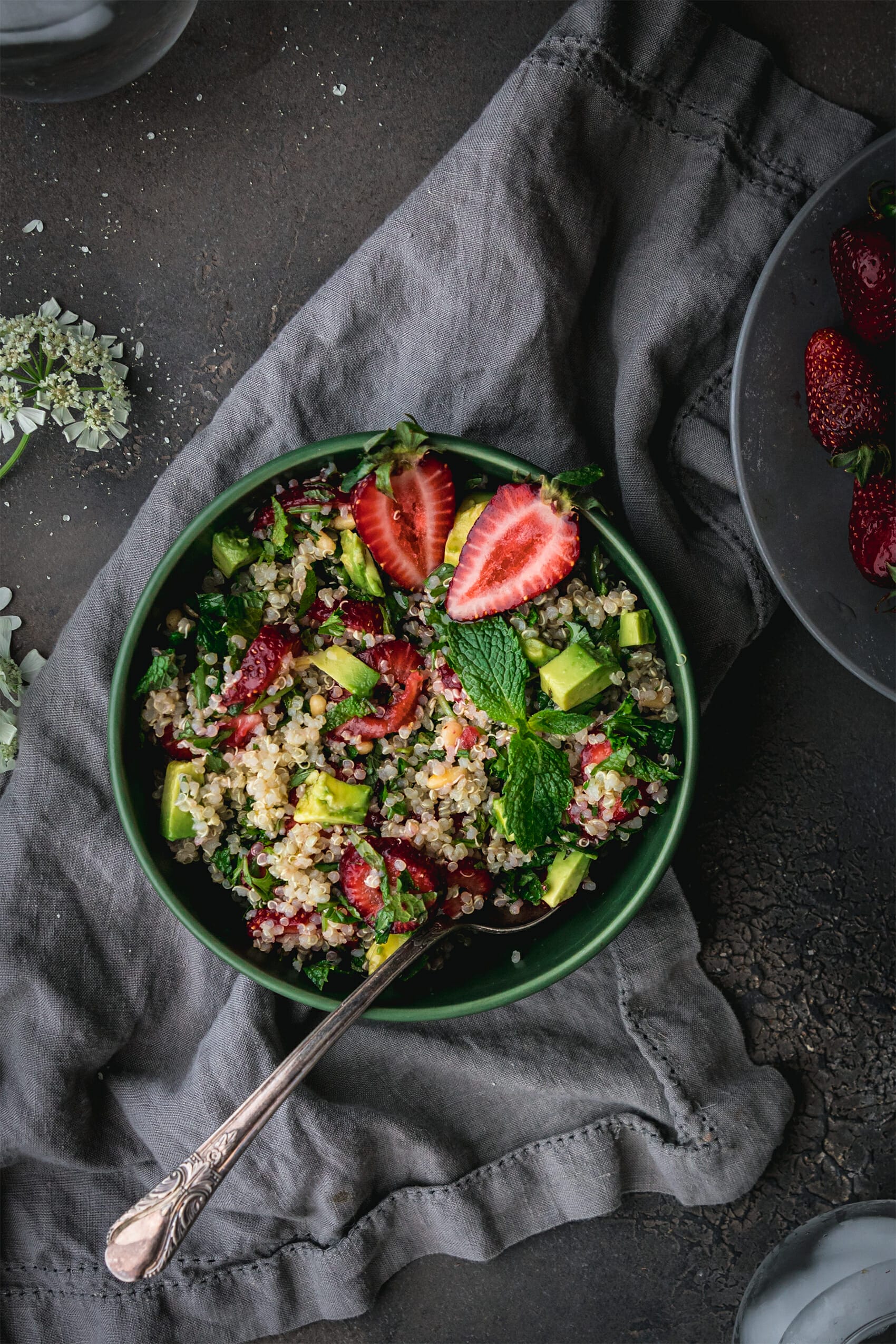 overhead view of green bowl of strawberry quinoa salad with a silver spoon on a grey table