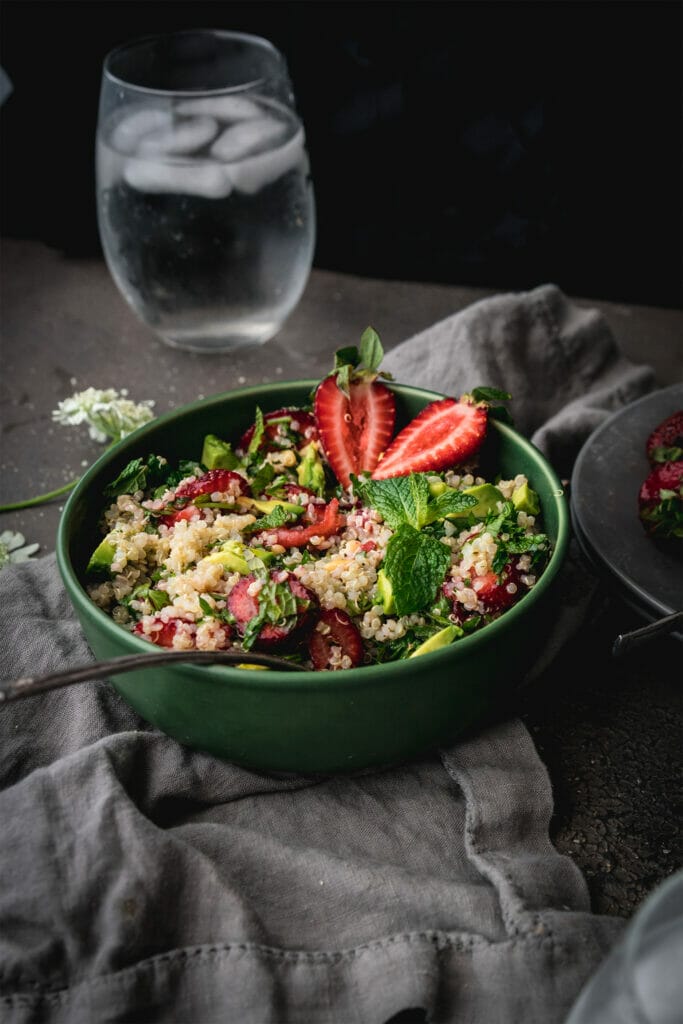 side view of green bowl of strawberry quinoa salad with a silver spoon on a grey table