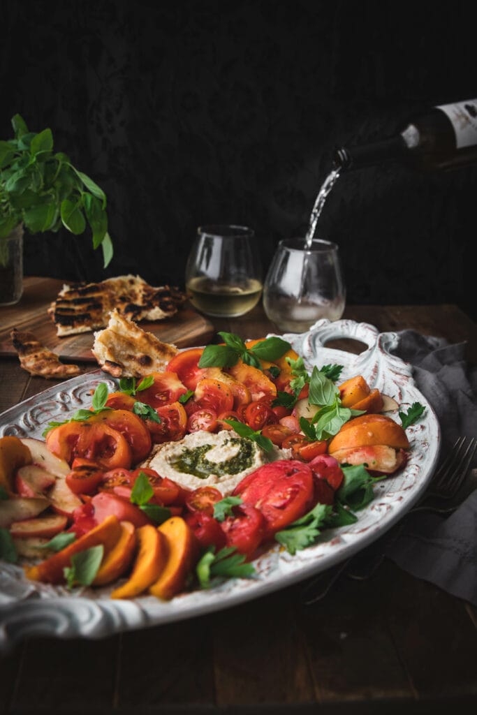 wine poured into a glass behind a tray of fresh tomatoes and sliced peaches in a caprese salad