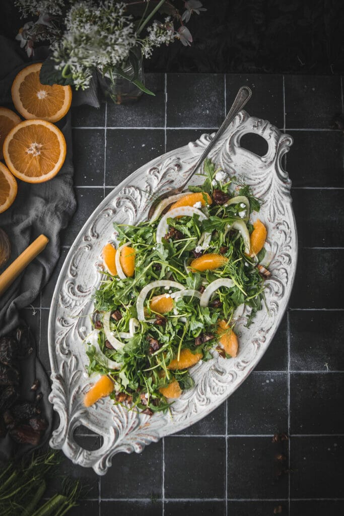 overhead shot of arugula fennel and orange salad on a white platter on a black table