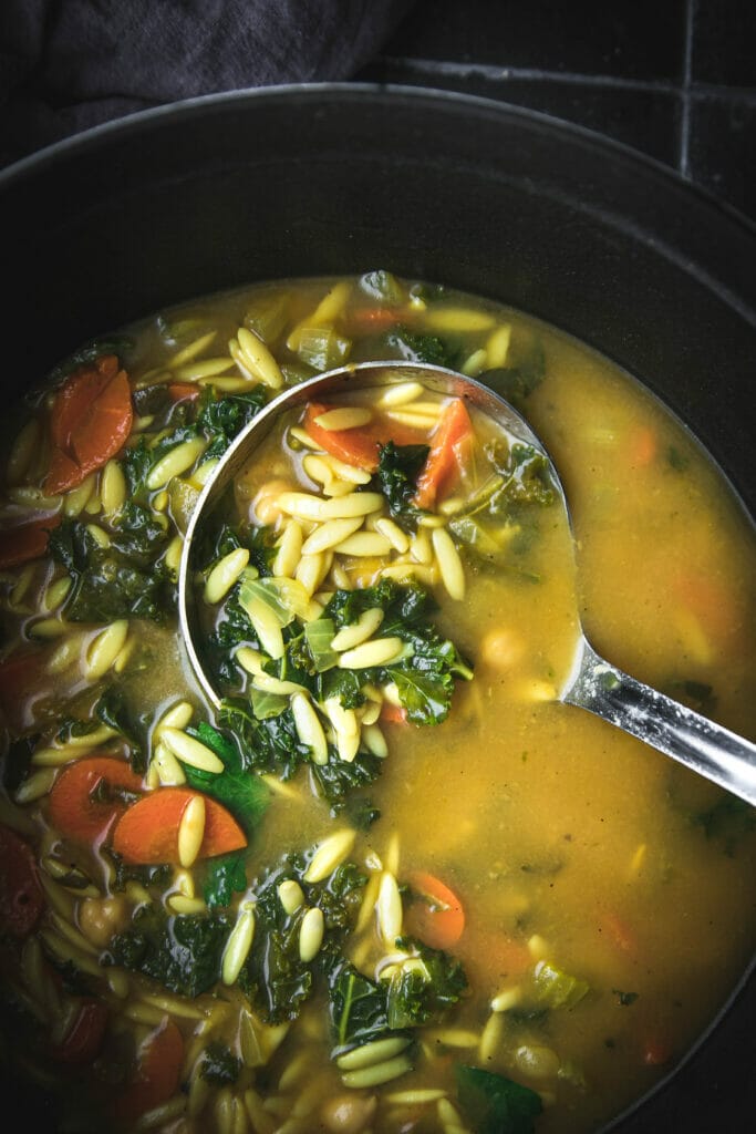 silver ladle in a pot of vegan chickpea orzo soup with kale
