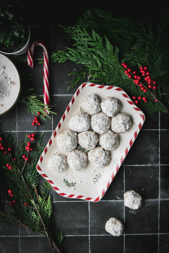 overhead shot of a tray of toasted walnut snowballs with powdered sugar on a red and white plate next to a candy cane