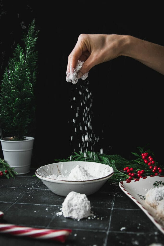 hand pouring powdered sugar onto a toasted walnut snowball cookie