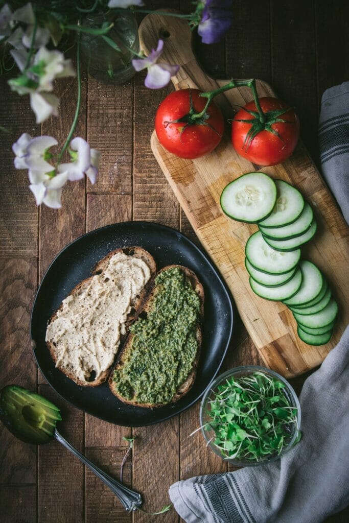 overhead shot of two slices of bread with hummus and pesto on a black plate next to cucumbers and tomatoes