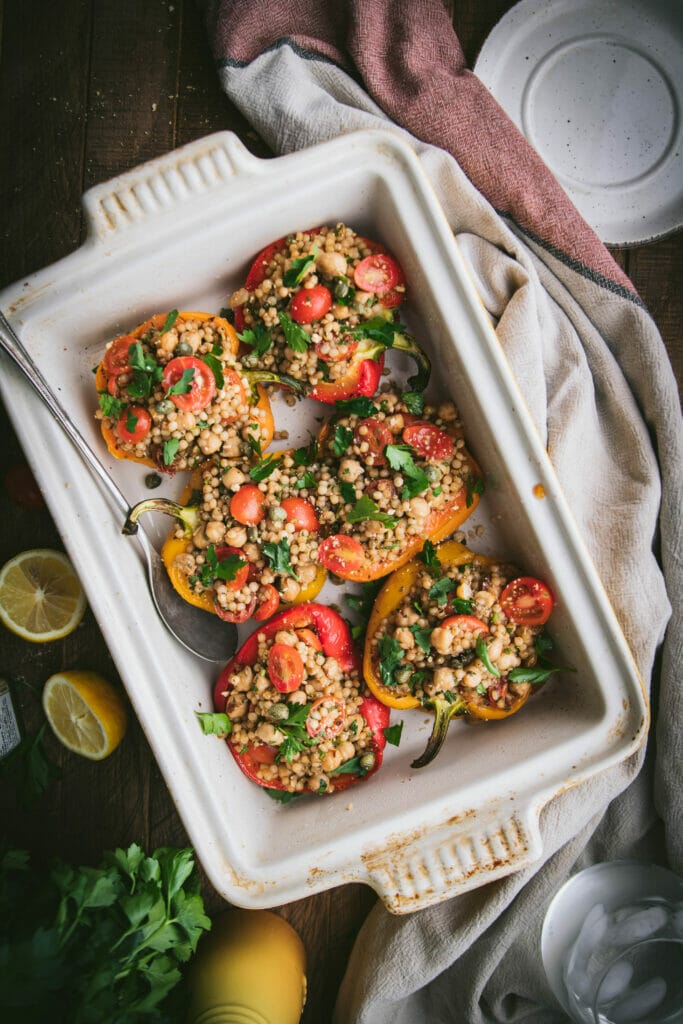 overhead shot of six vegan stuffed peppers with greek couscous in a rectangular baking dish next to a towel