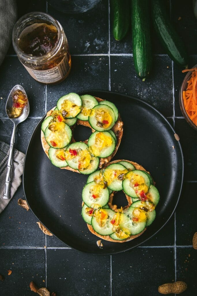 overhead shot of a sliced open thai peanut bagel with cucumber slices and jalapeño jam on a black plate