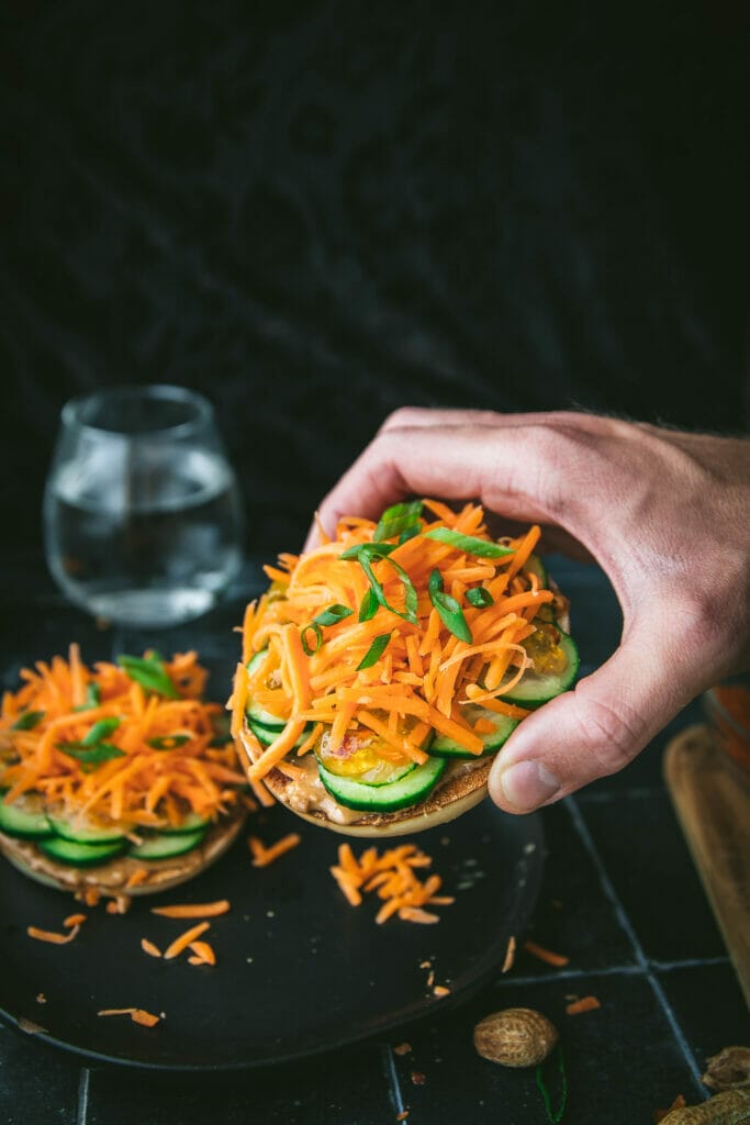 hand reaching for an open faced Thai peanut bagel with cucumber and carrot pieces