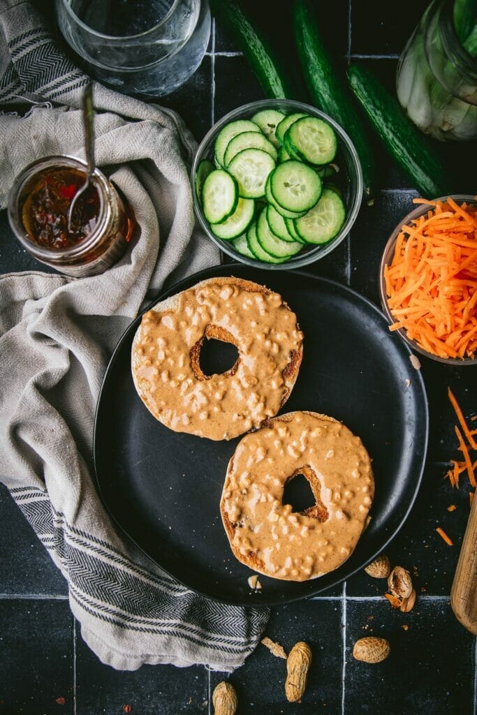 sliced bagel with peanut butter on a black plate next to cucumber and carrot pieces