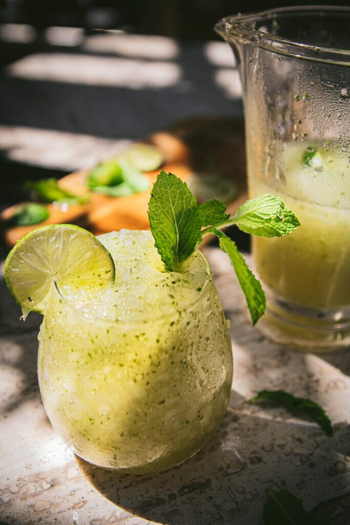 close up of a frozen mojito slushie in a glass with a lime and a mint sprig