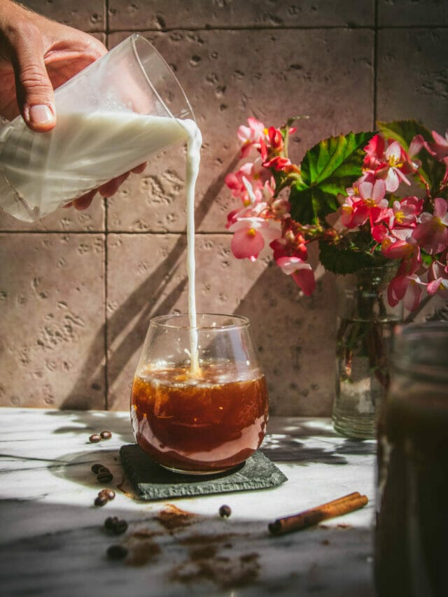 milk pouring into a glass of cinnamon cold brew next to a vase of flowers