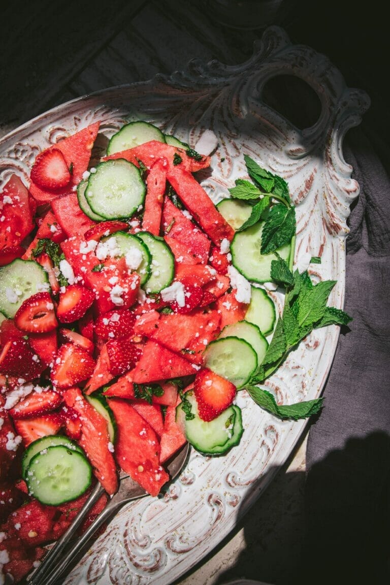 a high angle shot of watermelon, cucumber, and feta salad next to a bottle of water on a white tray