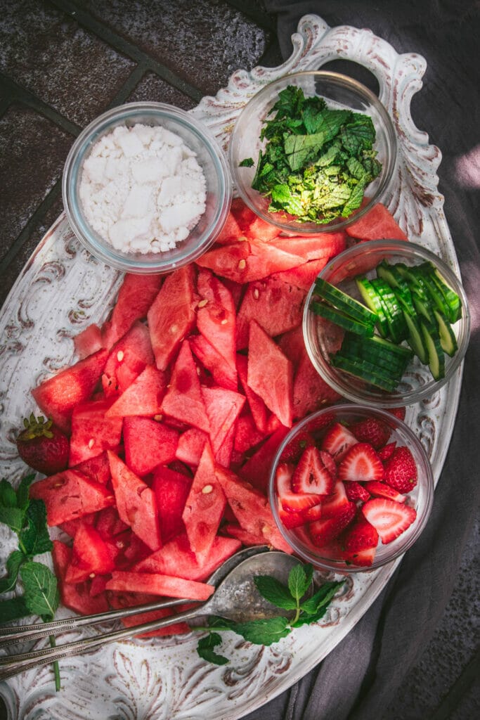 an overhead view of sliced watermelon with small bowls of mint, strawberries, cucumber, and feta