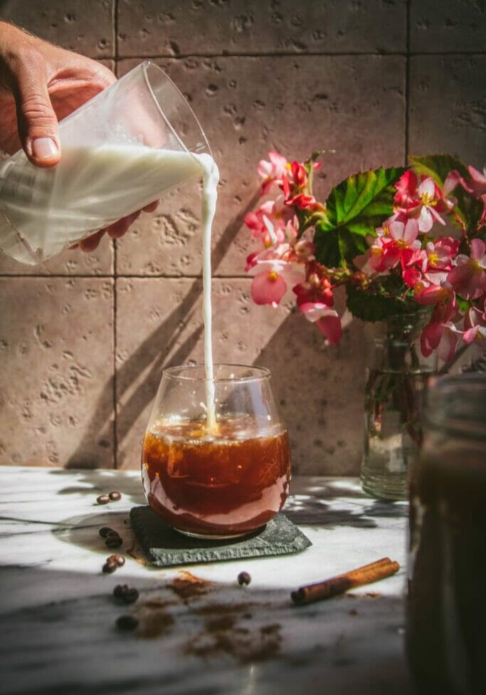 milk pouring into a glass of cinnamon cold brew next to a vase of flowers
