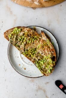 slice of tahini avocado toast on a white plate topped with old bay and everything bagel seasoning