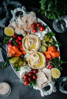 platter of creamy ottolenghi hummus surrounded by carrots, pickles, onions, pita, and cherry tomatoes with fresh herbs
