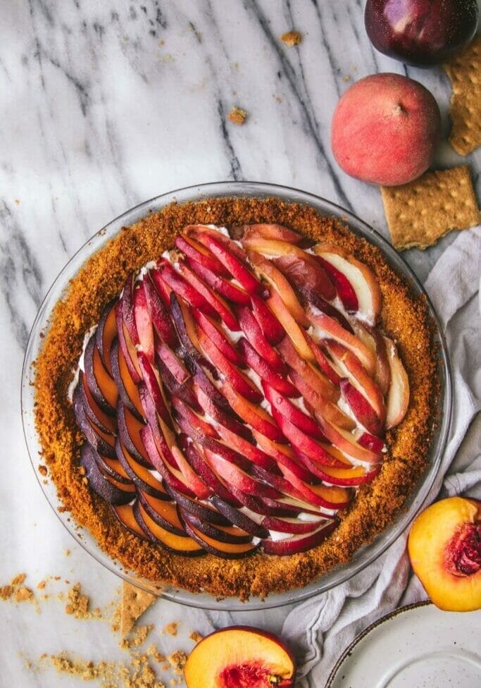 peaches and cream tart on a marble table next to sliced peaches and graham crackers