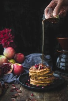 hand pouring maple syrup onto a stack of vegan pumpkin pancakes