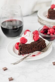 slice of red wine chocolate cake topped with raspberries and white whipped cream next to a stemless red wine glass