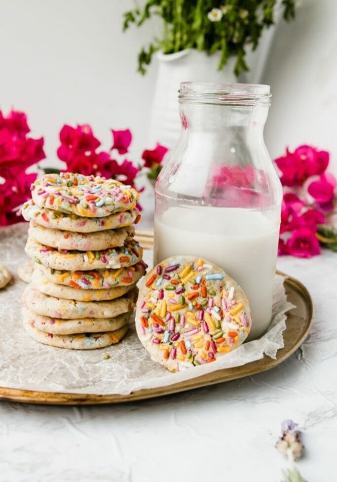 stack of rainbow sprinkle cookies on a tray next to milk